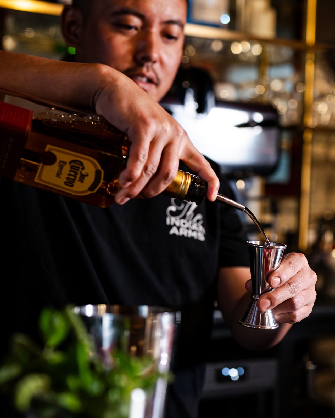 Barman en tenue noire versant un spiritueux dans un shaker au bar de The Indian Arms